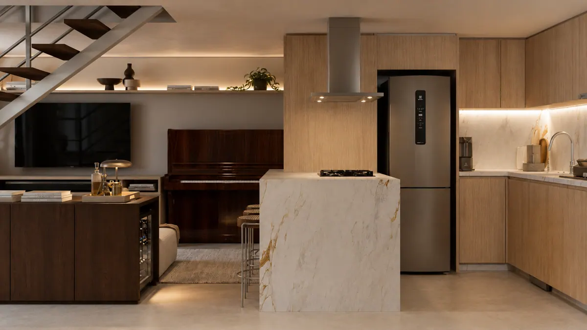 Cozinha em madeira clara com ilha de mármore e geladeira inox, com o piano e a TV ao lado
