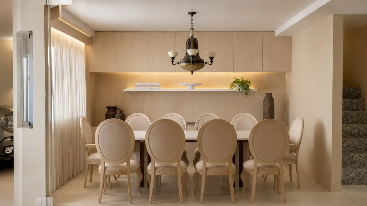 Sala de jantar de frente para a parede de marcenaria clara, com prateleira iluminada e lustre clássico