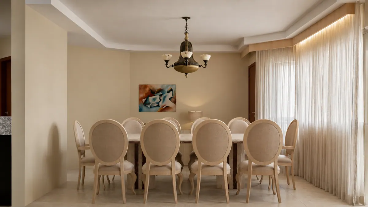 Sala de jantar vista da cabeceira, com quadro abstrato, abajur e cortinas de linho banhadas de luz